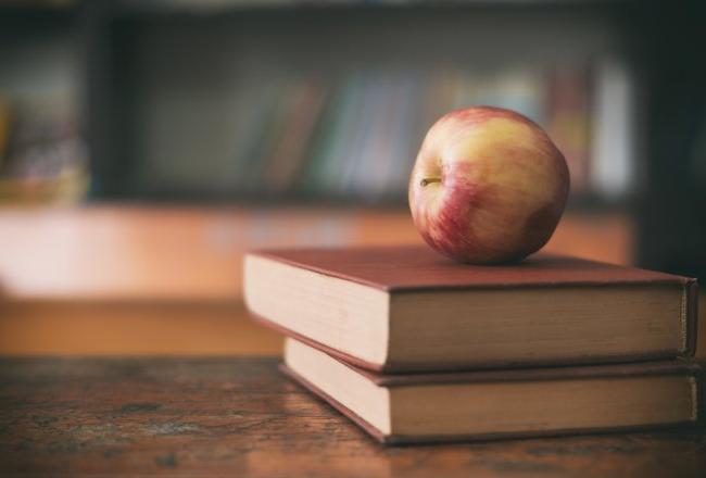 Apple sitting on a pile of books on a classroom desk.